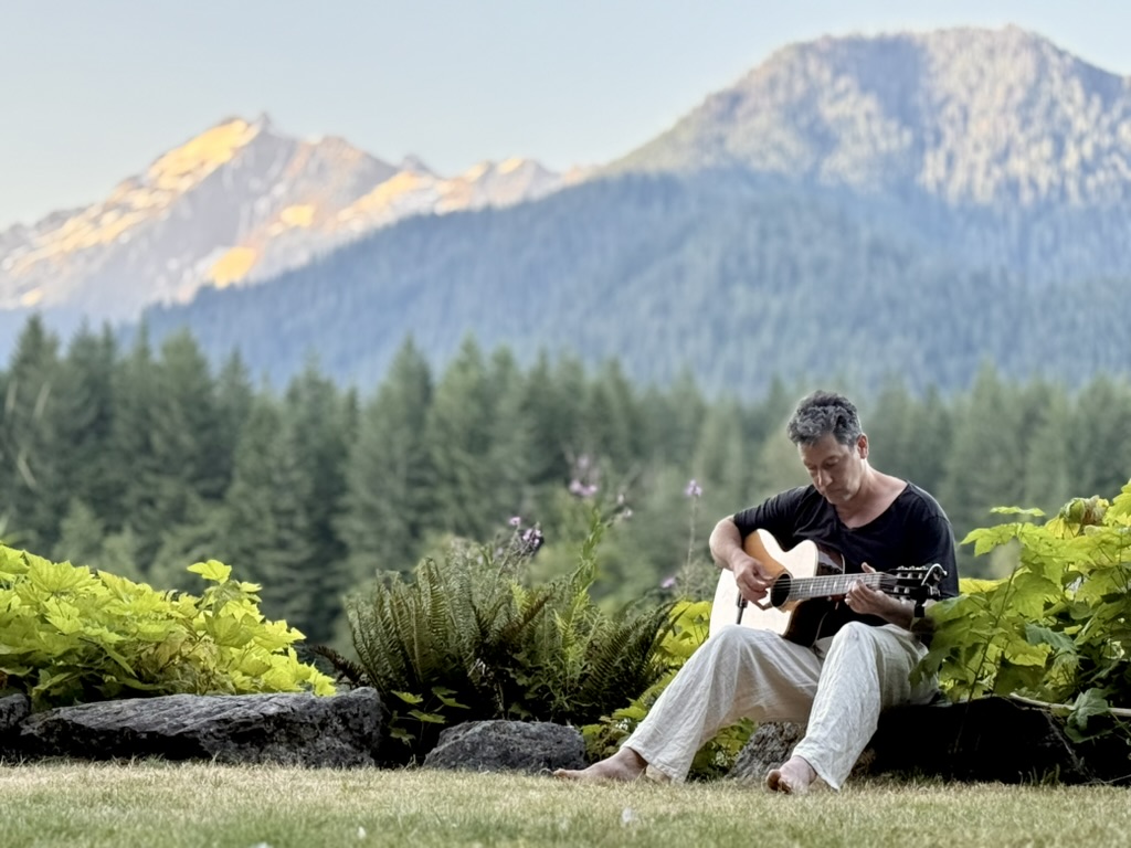 Chris Weber playing guitar among ferns with mountains behind, golden hour light on peaks